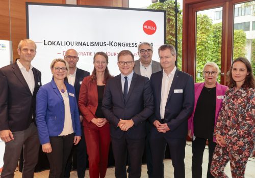 Vier Frauen und fünf Männer stehen vor einem Lokaljournalismus-Kongress Banner und schauen in die Kamera