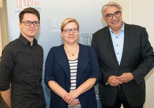 Zwei Männer und eine Frau stehen nebeneinander vor einem Banner mit der Aufschrift TLM