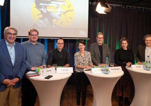 Fünf Männer und zwei Frauen stehen nebeneinander an Stehtischen vor einer Beamerleinwand in einem Studio