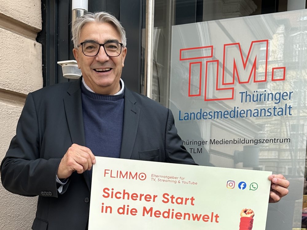 Ein Mann mit grauen Haaren und Brille hält ein FLIMMO-Plakat für die Kindertagesstätten in die Kamera