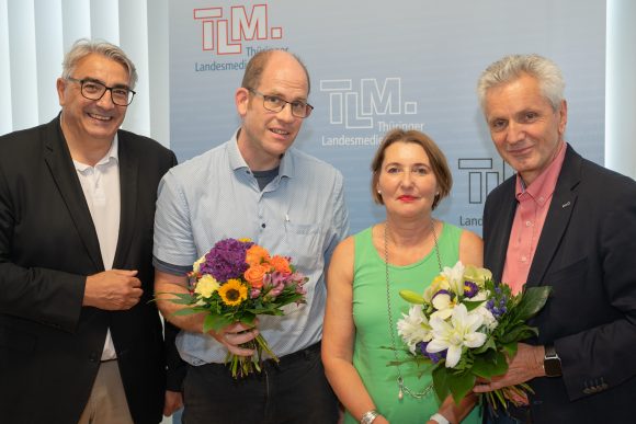 Drei Männer und eine Frau stehen vor einem TLM-Banner und lächeln in die Kamera. Zwei Männer halten einen Blumenstrauß.