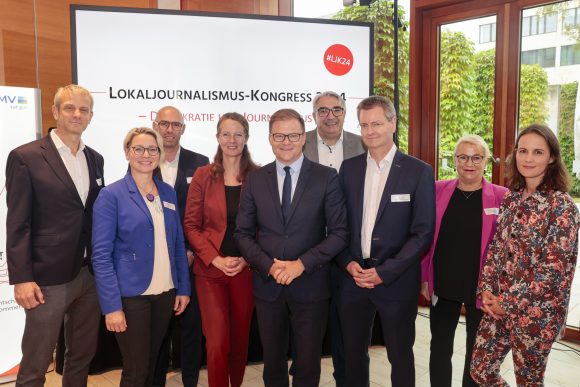 Vier Frauen und fünf Männer stehen vor einem Lokaljournalismus-Kongress Banner und schauen in die Kamera