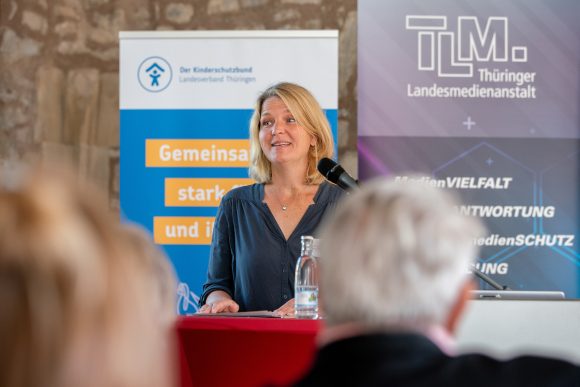 Eine blonde Frau steht an einem Rednerpult. Im Hintergrund stehen Banner der TLM und des Kinderschutzbundes.