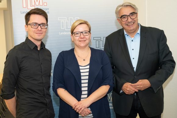 Zwei Männer und eine Frau stehen nebeneinander vor einem Banner mit der Aufschrift TLM