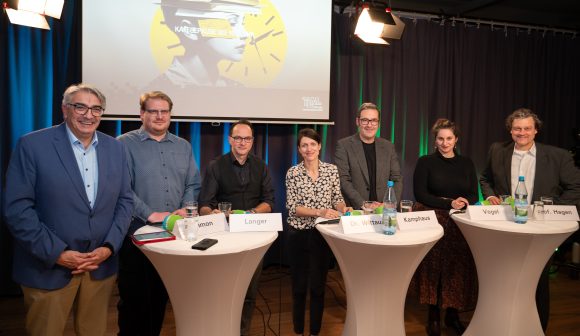 Fünf Männer und zwei Frauen stehen nebeneinander an Stehtischen vor einer Beamerleinwand in einem Studio